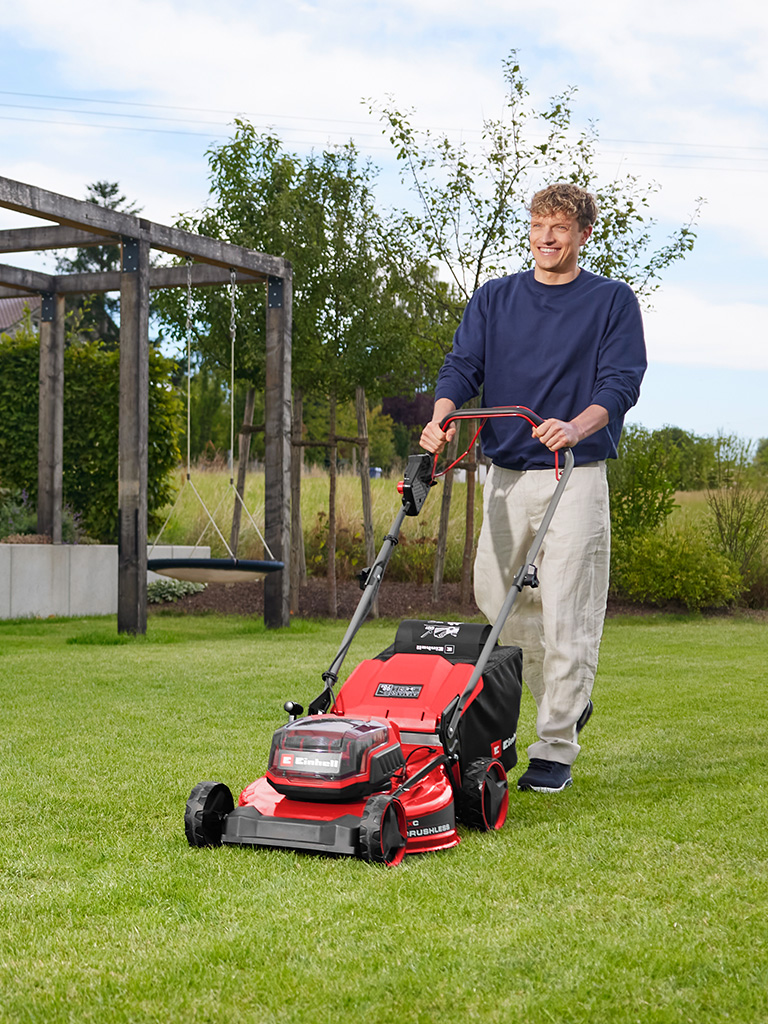 Ein Mann mäht eine Rasenfläche mit dem roten Einhell Akku-Rasenmäher GC-CM 36/51 S Li BL vor einer Gartenanlage mit Holzpergola.