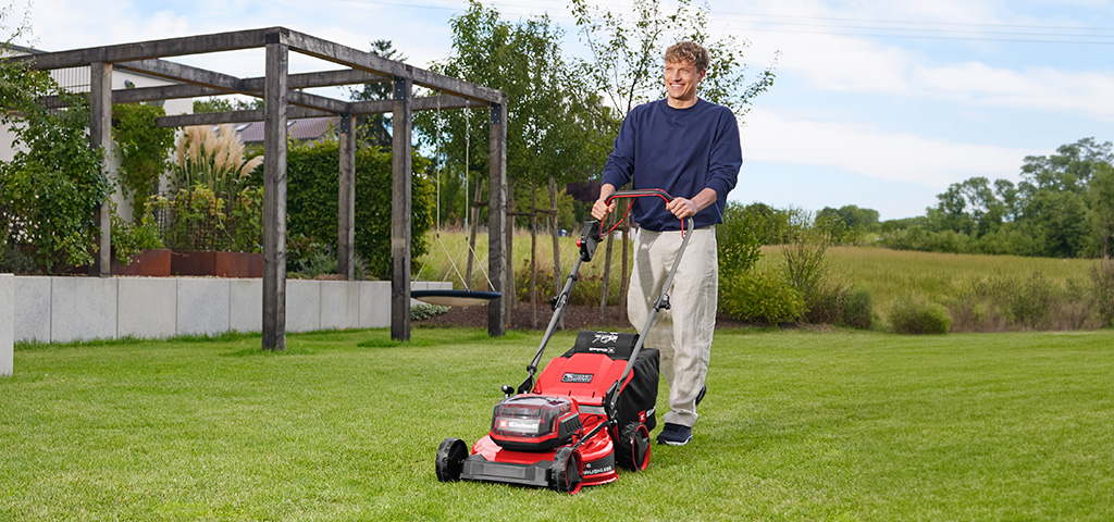 Ein Mann mäht eine Rasenfläche mit dem roten Einhell Akku-Rasenmäher GC-CM 36/51 S Li BL vor einer Gartenanlage mit Holzpergola.