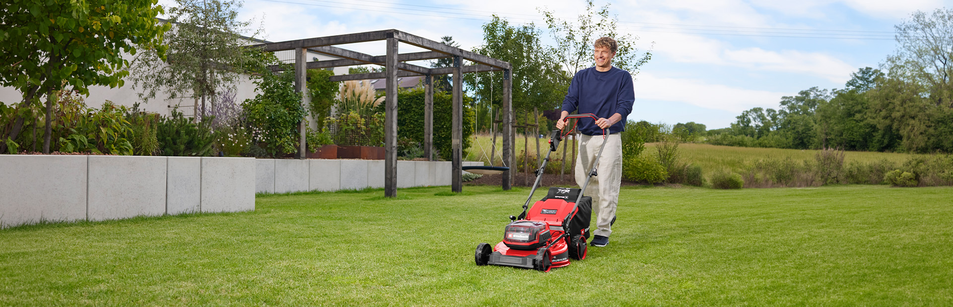 Ein Mann mäht eine Rasenfläche mit dem roten Einhell Akku-Rasenmäher GC-CM 36/51 S Li BL vor einer Gartenanlage mit Holzpergola.