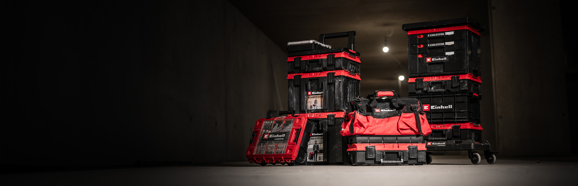 Mehrere rote Einhell E-Case Werkzeugkoffer mit schwarzen Details, teils gestapelt, sowie eine rote Werkzeugtasche in einer dunklen Werkstatt.