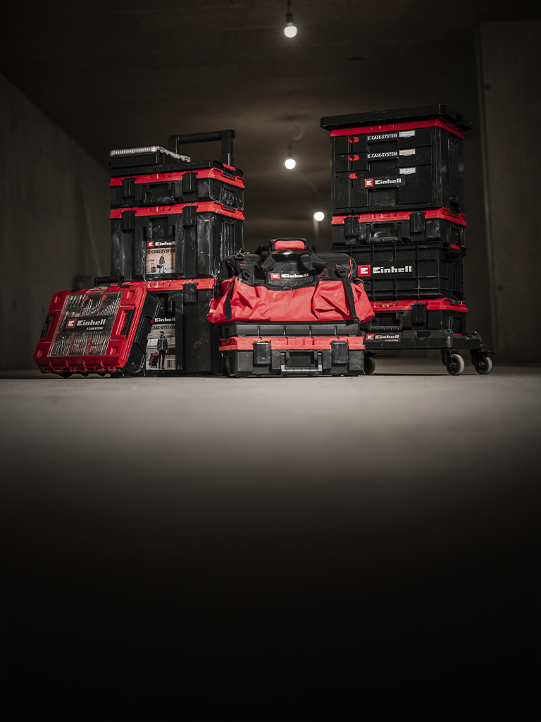 Mehrere rote Einhell E-Case Werkzeugkoffer mit schwarzen Details, teils gestapelt, sowie eine rote Werkzeugtasche in einer dunklen Werkstatt.