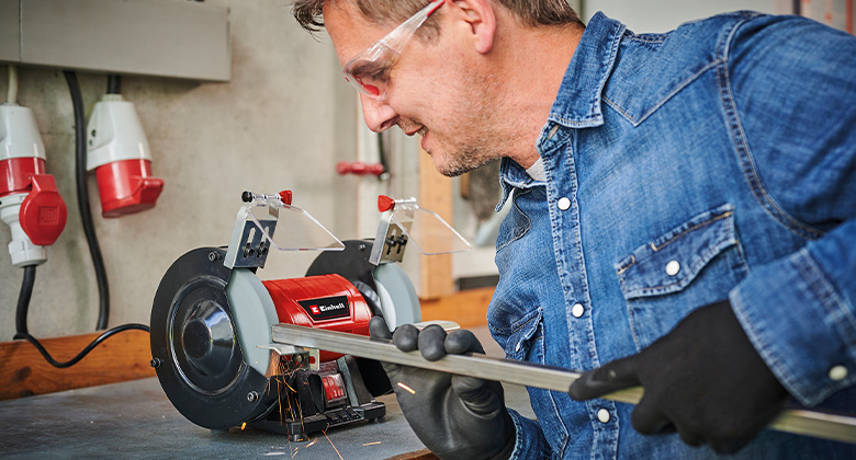 Ein Mann schleift eine Metallstange mit einem Einhell Doppelschleifer in einer Werkstatt.