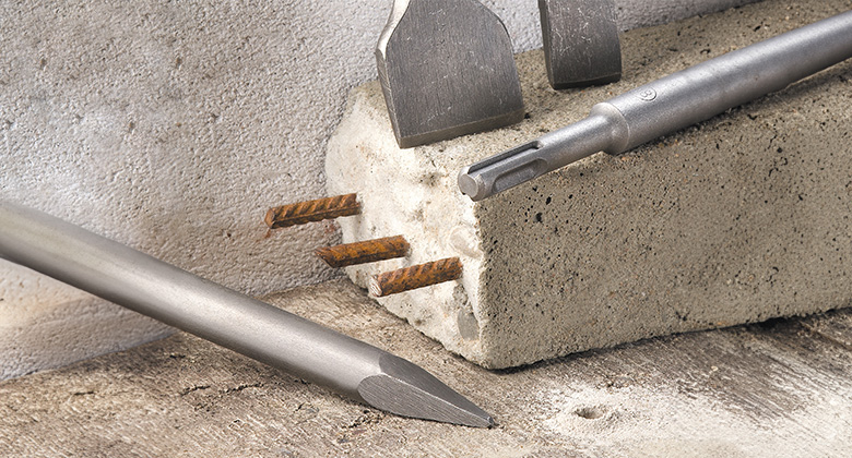 Verschiedene Meißelaufsätze liegen neben einem Betonblock mit freiliegenden Metallstäben.