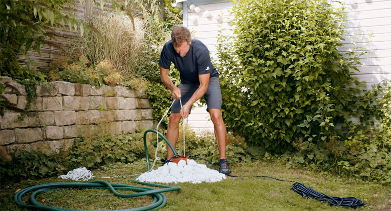 Ein Mann steht über einem Rohr gebeugt im Garten, in der Hand ein Seil, an dem eine Tiefbrunnenpumpe befestigt ist.