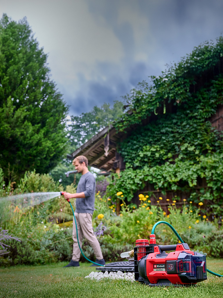 Ein Mann bewässert mit der Einhell Akku-Gartenpumpe AQUINNA im Garten Blumenbeete mit einem Gartenschlauch.
