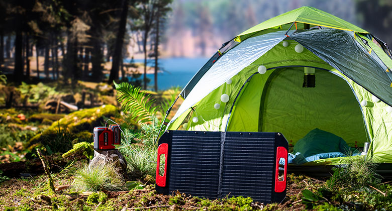 Green camping tent in a forest with a set-up solar panel and a small portable generator on the ground.