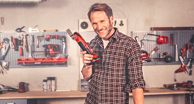 Ein Mann steht in seiner Werkstatt und hält ein kabelloses Multifunktionswerkzeug von Einhell in der Hand.