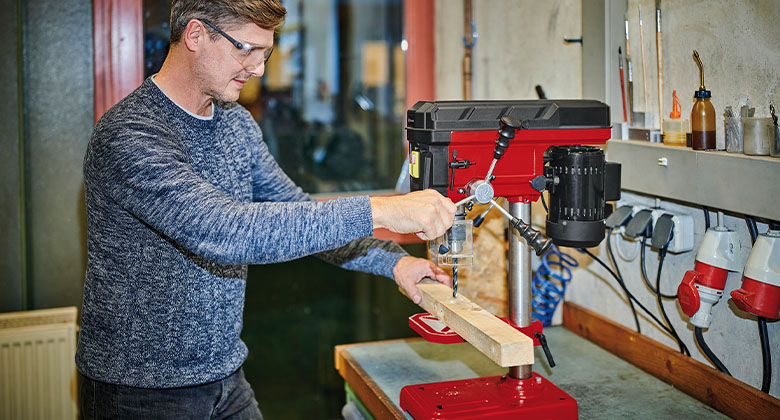 Ein Mann bohrt mit einer Tischbohrmaschine in eine Holzlatte.