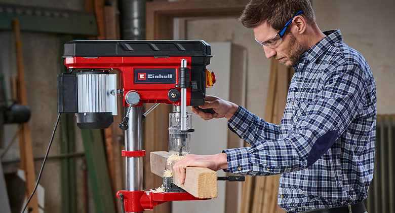 Ein Mann mit Schutzbrille bohrt mit Hilfe einer Säulenbohrmaschine ein Loch in eine Holzlatte.