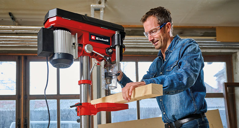 Ein Mann nutzt eine Tischbohrmaschine, um ein Loch in eine dicke Holzlatte zu bohren.