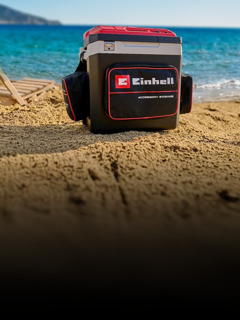 Das Bild zeigt eine Einhell-Kühlbox am Strand neben einer Liege. Im Hintergrund sieht man das blaue Meer und eine Küstenlandschaft.