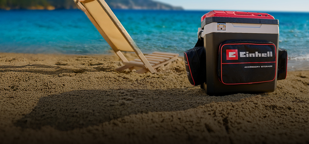 Das Bild zeigt eine Einhell-Kühlbox am Strand neben einer Liege. Im Hintergrund sieht man das blaue Meer und eine Küstenlandschaft.