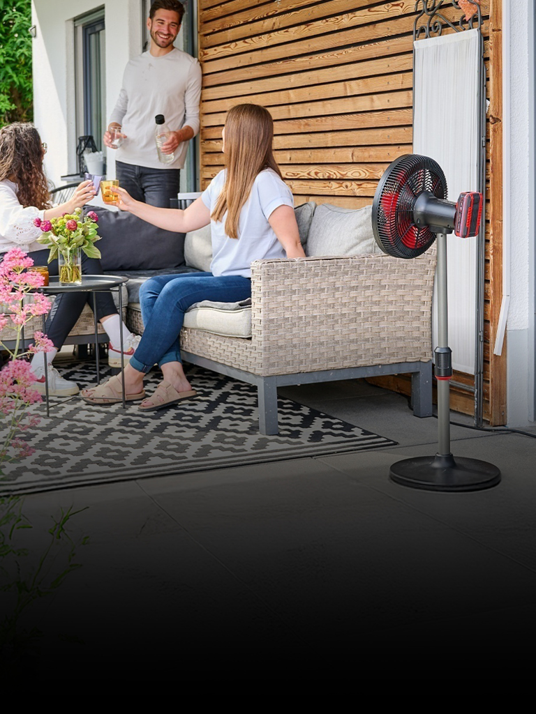 Drei Personen genießen Getränke auf einer gemütlichen Terrasse, umgeben von Blumen und Grünpflanzen. Ein rot-schwarzer Einhell-Akku-Ventilator steht neben der Sitzecke und sorgt an einem sonnigen Tag für frische Luft.