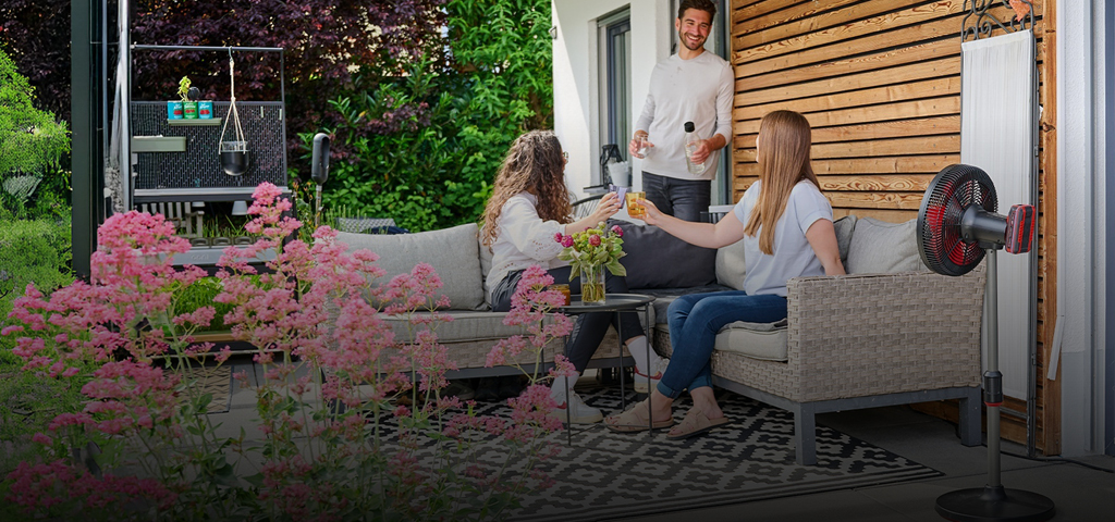 Drei Personen genießen Getränke auf einer gemütlichen Terrasse, umgeben von Blumen und Grünpflanzen. Ein rot-schwarzer Einhell-Akku-Ventilator steht neben der Sitzecke und sorgt an einem sonnigen Tag für frische Luft.