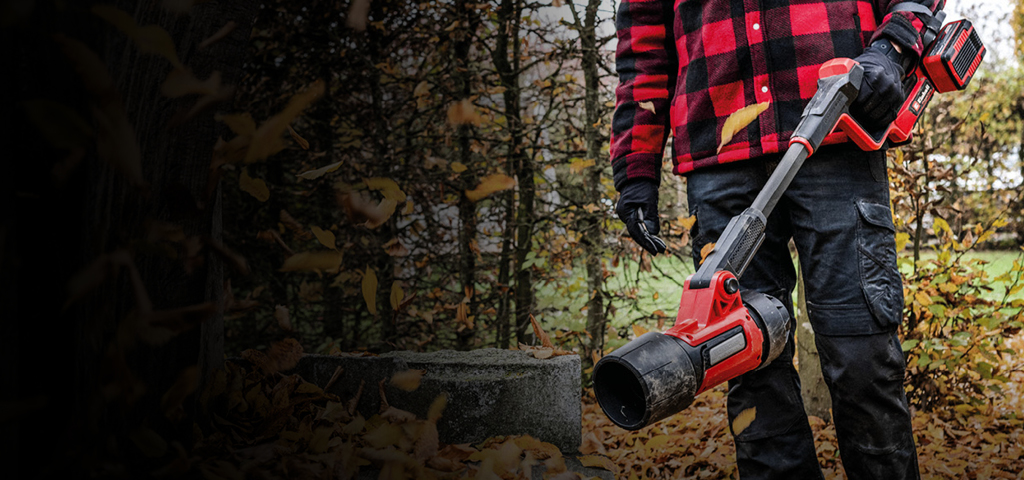 ChatGPT:  A person wearing black pants, gloves, and a red-and-black checkered jacket is standing outdoors holding a red cordless leaf blower from Einhell. Fallen autumn leaves cover the ground, and trees can be seen in the background.
