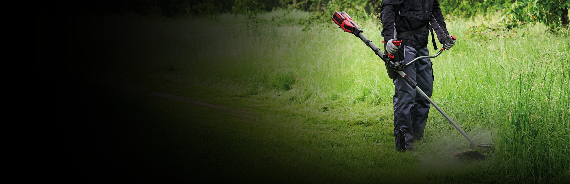 A man in protective gear, including a red helmet and ear protection, uses a red and black Einhell cordless brush cutter to trim tall grass along a path in a green, wooded area.