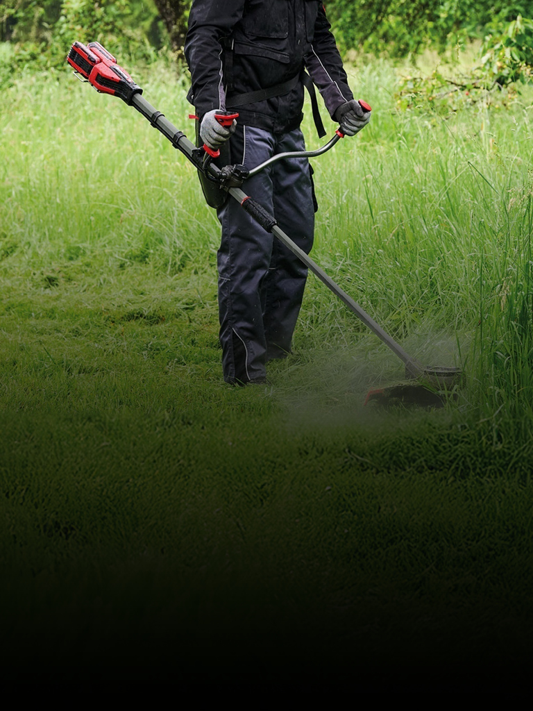 A man in protective gear, including a red helmet and ear protection, uses a red and black Einhell cordless brush cutter to trim tall grass along a path in a green, wooded area.