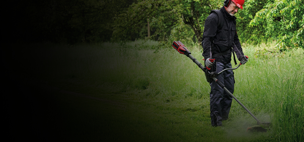 A man in protective gear, including a red helmet and ear protection, uses a red and black Einhell cordless brush cutter to trim tall grass along a path in a green, wooded area.