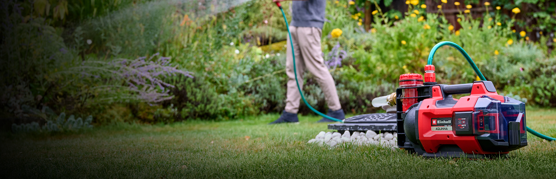 Ein Mann in Freizeitkleidung bewässert seinen Garten mit einem Schlauch, der mit einer roten und schwarzen Einhell-Akku-Wasserpumpe verbunden ist, die im Vordergrund auf dem Rasen steht. Im Hintergrund sind üppige grüne Pflanzen und gelbe Blumen zu sehen.