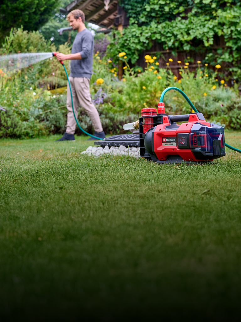 Ein Mann in Freizeitkleidung bewässert seinen Garten mit einem Schlauch, der mit einer roten und schwarzen Einhell-Akku-Wasserpumpe verbunden ist, die im Vordergrund auf dem Rasen steht. Im Hintergrund sind üppige grüne Pflanzen und gelbe Blumen zu sehen.