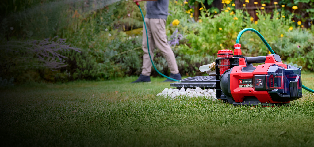 Ein Mann in Freizeitkleidung bewässert seinen Garten mit einem Schlauch, der mit einer roten und schwarzen Einhell-Akku-Wasserpumpe verbunden ist, die im Vordergrund auf dem Rasen steht. Im Hintergrund sind üppige grüne Pflanzen und gelbe Blumen zu sehen.