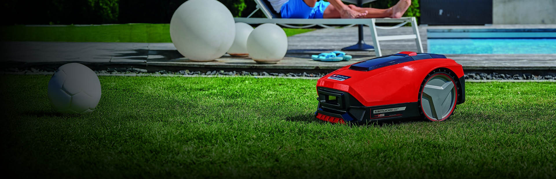 Ein Mann entspannt sich auf einer Sonnenliege in der Nähe eines Pools, während im Vordergrund ein roter Mähroboter selbstständig den Rasen trimmt.