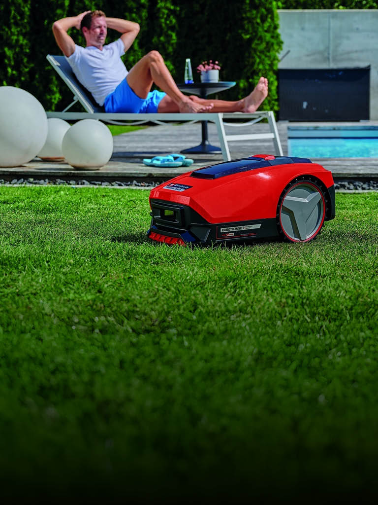 Ein Mann entspannt sich auf einer Sonnenliege in der Nähe eines Pools, während im Vordergrund ein roter Mähroboter selbstständig den Rasen trimmt.