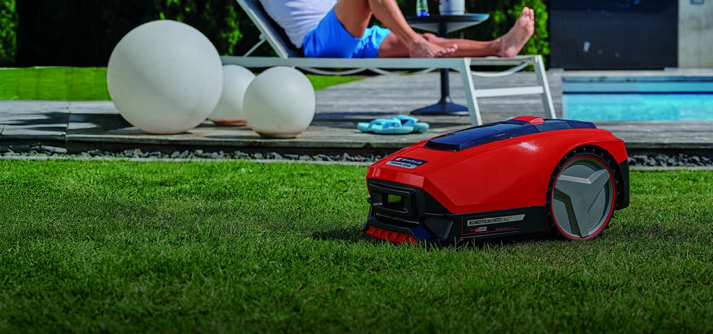 Ein Mann entspannt sich auf einer Sonnenliege in der Nähe eines Pools, während im Vordergrund ein roter Mähroboter selbstständig den Rasen trimmt.