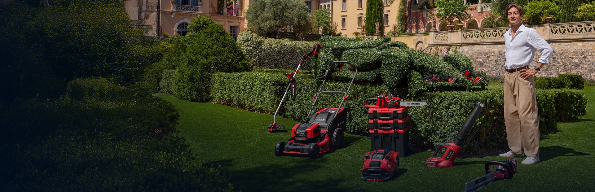 George Russell stands in a Mediterranean garden with Einhell cordless garden tools. Next to him, a race car-shaped hedge. 