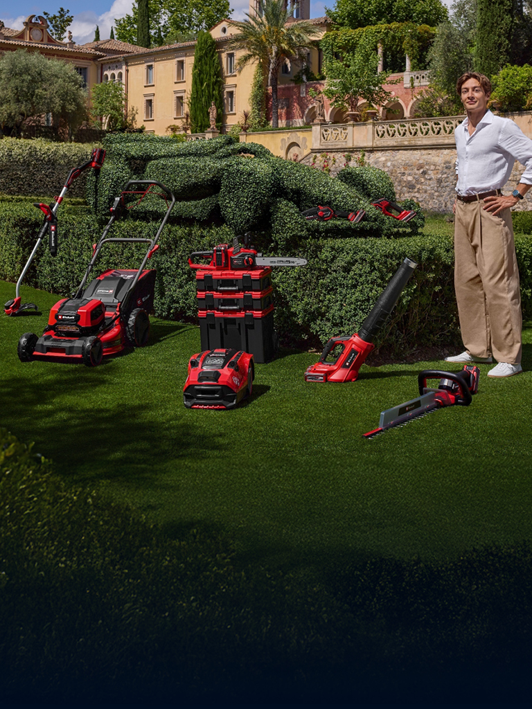 George Russell stands in a Mediterranean garden with Einhell cordless garden tools. Next to him, a race car-shaped hedge. 