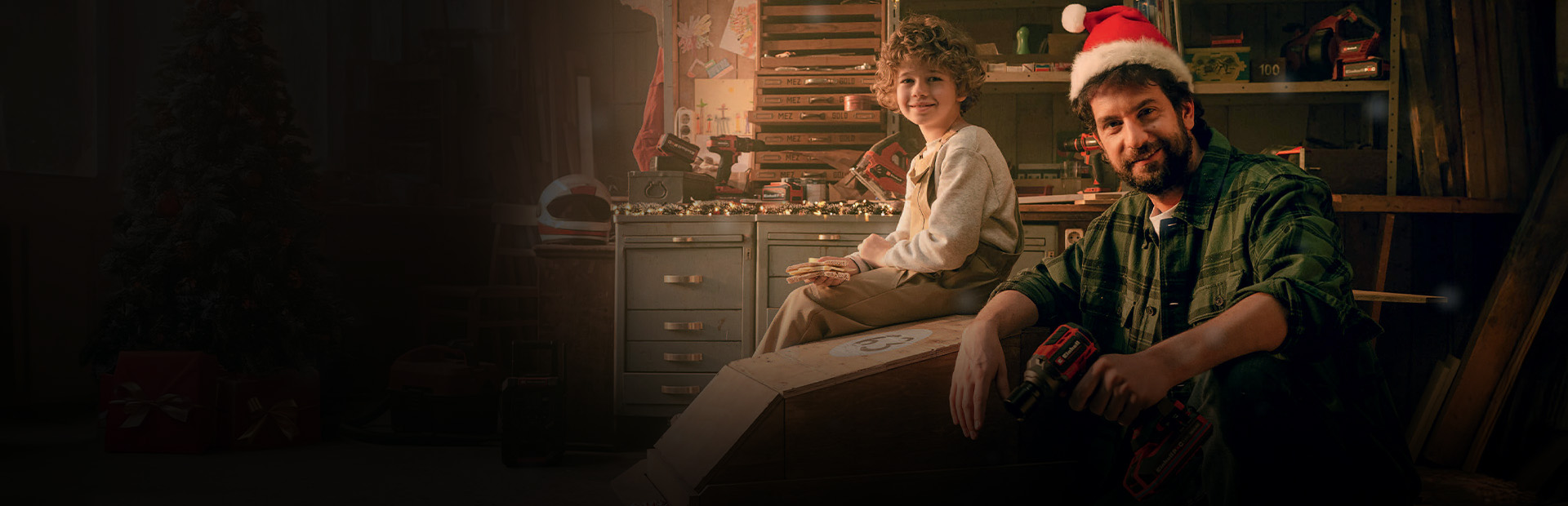 Ein Kind sitzt auf einem Holzauto, ein Mann mit Weihnachtsmütze kniet daneben mit einem Akkuschrauber. Im Hintergrund ist eine Werkbank, daneben ein Geschenk in rotem Papier.