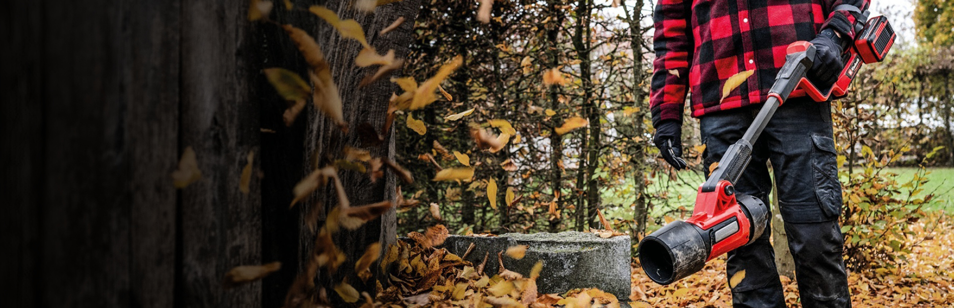 A person wearing black pants, gloves, and a red-and-black checkered jacket is standing outdoors holding a red cordless leaf blower from Einhell. Fallen autumn leaves cover the ground, and trees can be seen in the background.