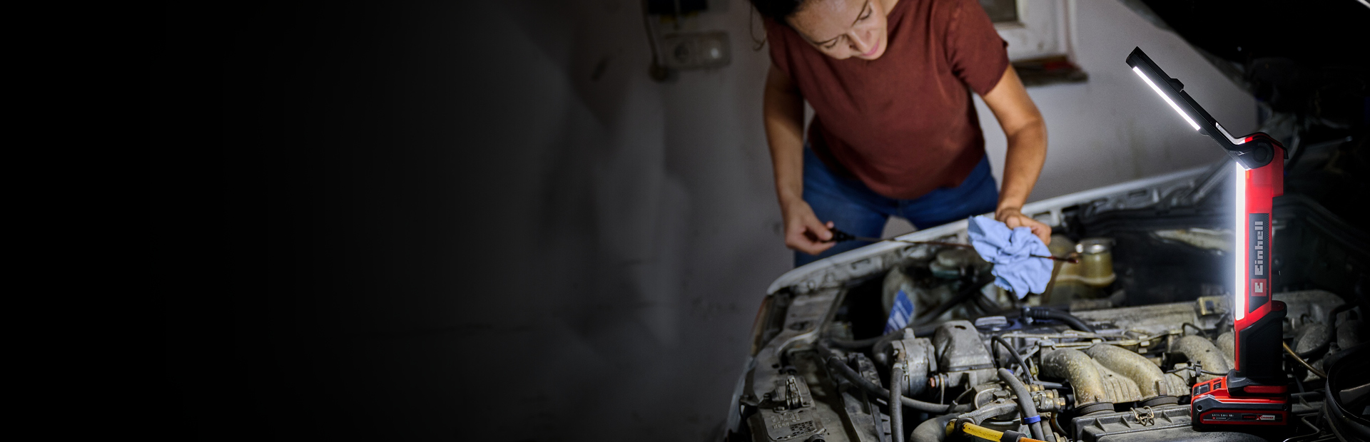 The image shows a person working on a car engine in a dimly lit garage. A bright red and black Einhell work light illuminates the engine area, providing clear visibility while the person uses a cloth and tools.