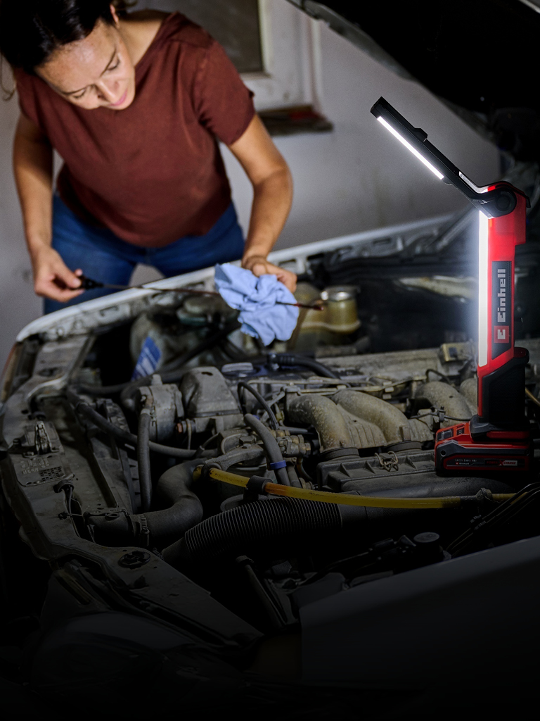 The image shows a person working on a car engine in a dimly lit garage. A bright red and black Einhell work light illuminates the engine area, providing clear visibility while the person uses a cloth and tools.