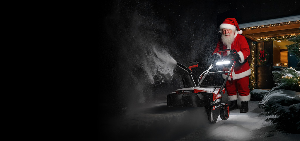 Eine als Weihnachtsmann verkleidete Person steht im Schnee vor einem mit Weihnachtsbeleuchtung geschmückten Haus und benutzt eine Schneefräse.