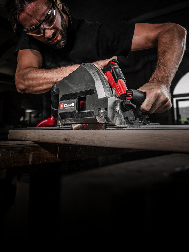 Ein Mann mit Schutzbrille bedient eine leistungsstarke Einhell-Handkreissäge, um ein Holzbrett in einer Werkstatt zu schneiden. Sein konzentrierter Gesichtsausdruck und fester Griff betonen Präzision und Sicherheit.