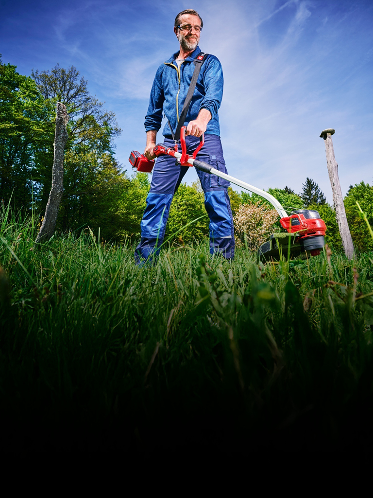Ein Mann in blauer Arbeitskleidung und mit Schutzbrille trimmt mit einem rot-schwarzen Akku-Rasentrimmer von Einhell hohes Gras zwischen Baumstümpfen in einer grünen, bewaldeten Gegend.