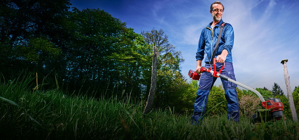 Ein Mann in blauer Arbeitskleidung und mit Schutzbrille trimmt mit einem rot-schwarzen Akku-Rasentrimmer von Einhell hohes Gras zwischen Baumstümpfen in einer grünen, bewaldeten Gegend.