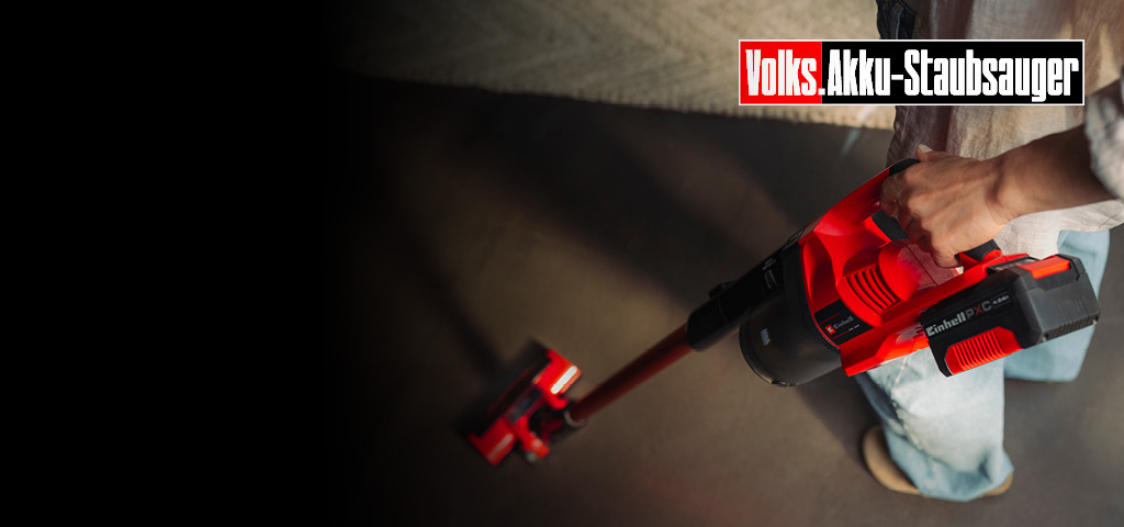Das Bild zeigt eine Person, die einen roten kabellosen Stabstaubsauger auf einem Boden benutzt. Sichtbares Branding und Text lauten „Volks Akku‑Staubsauger“ und „Einhell“, was auf einen akkubetriebenen Staubsauger hinweist.