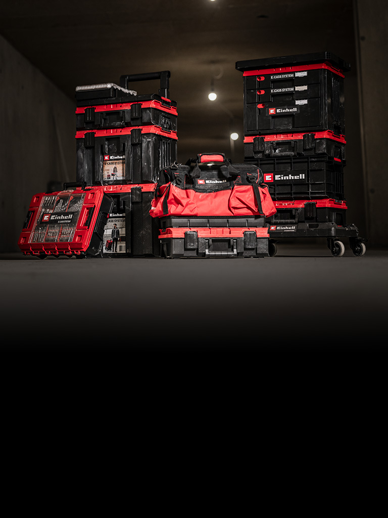 Der Banner zeigt einen Satz gestapelter Einhell E-Cases und eine rot-schwarze Werkzeugtasche, die auf einem Rollwagen angeordnet sind. Sie befinden sich in einer dunklen, industriell wirkenden Umgebung mit gezielter Beleuchtung, die die Produkte hervorhebt.