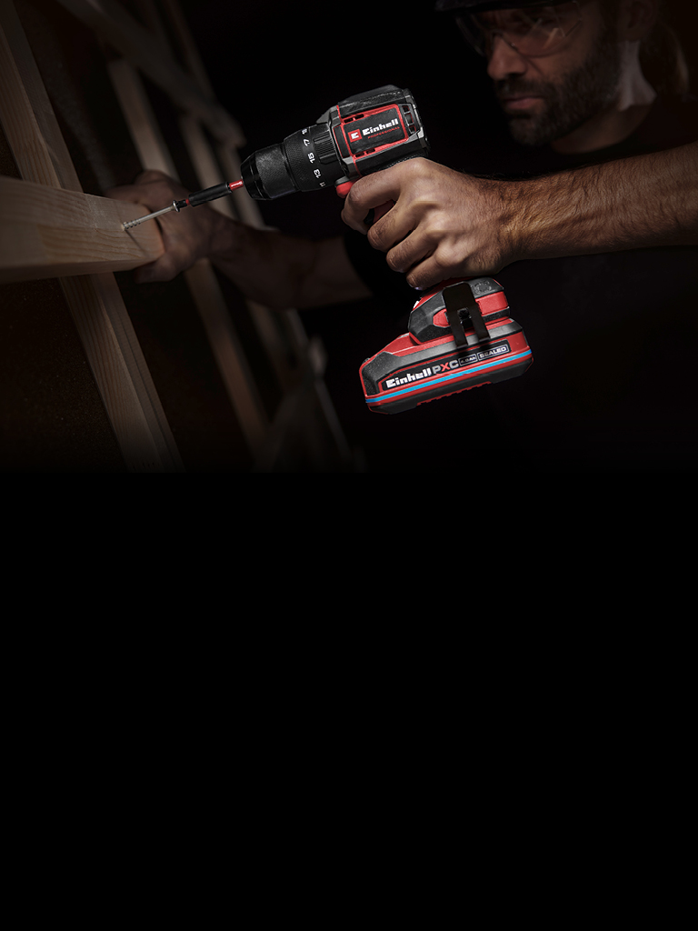 Ein Mann verwendet einen Akku-Schlagbohrschrauber, um eine Schraube in ein Stück Holz zu drehen. Die Szene ist dunkel, nur das Werkzeug und seine Hände sind im Vordergrund beleuchtet.