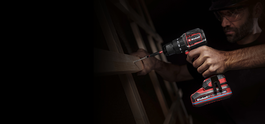 Ein Mann verwendet einen Akku-Schlagbohrschrauber, um eine Schraube in ein Stück Holz zu drehen. Die Szene ist dunkel, nur das Werkzeug und seine Hände sind im Vordergrund beleuchtet.