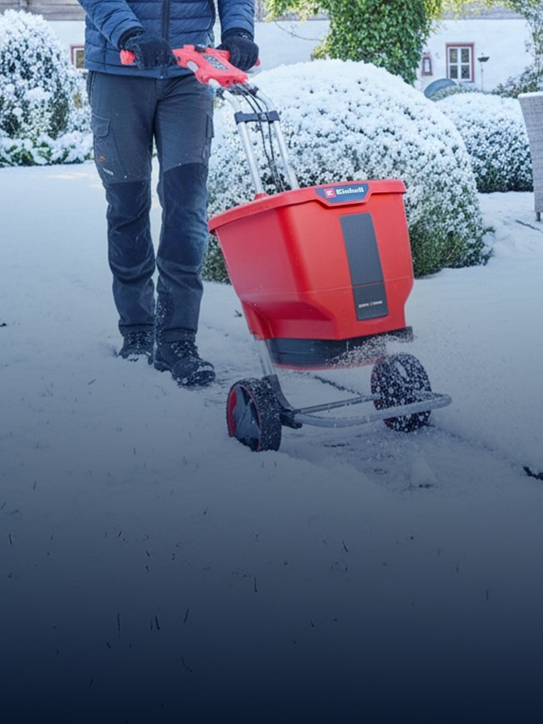 Eine Person benutzt einen roten Akku-Streuwagen auf einer schneebedeckten Gartenfläche in der Nähe eines Hauses und von Terrassenmöbeln. Der Boden, die Büsche und die Umgebung sind mit Schnee bedeckt.