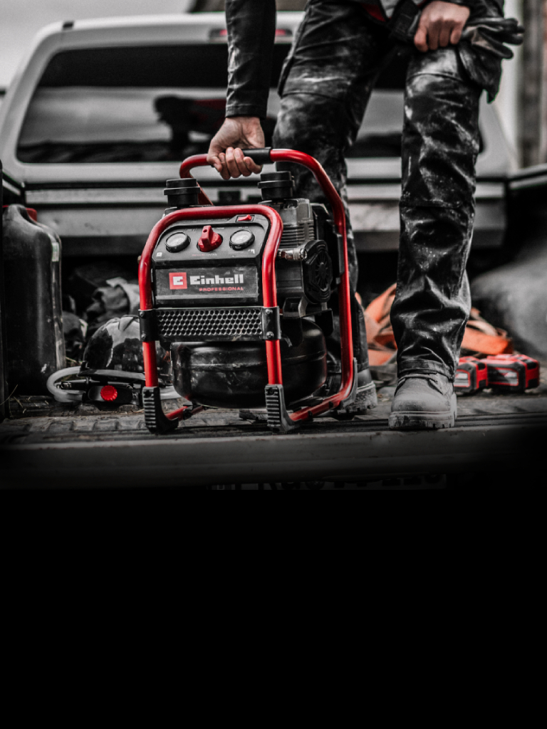 Auf dem Bild steht ein tragbarer Einhell‑Kompressor auf der offenen Ladefläche eines Fahrzeugs und wird von einer Person am Griff gehalten. Die Szene wirkt wie eine Arbeitssituation, wobei der Kompressor farblich hervorsticht.