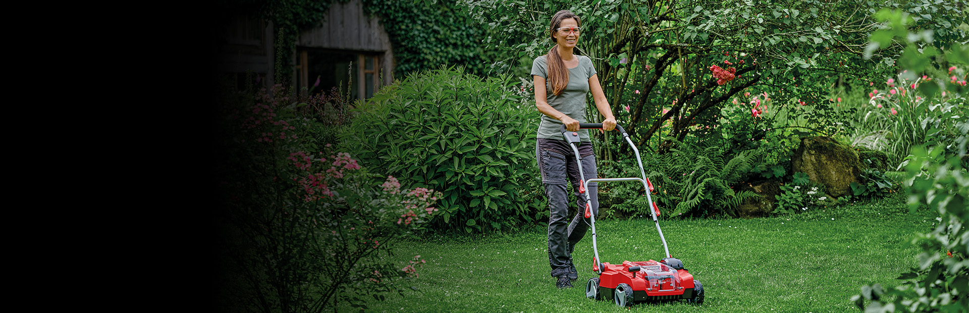 Die Aufnahme zeigt eine Person, die einen roten Rasenmäher über eine gepflegte, grüne Rasenfläche in einem üppigen Garten schiebt. Umgeben ist die Szene von dichtem Strauchwerk, Blumen und Bäumen.