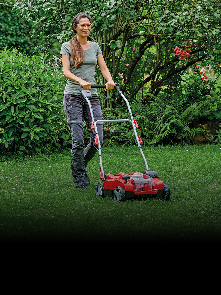 Die Aufnahme zeigt eine Person, die einen roten Rasenmäher über eine gepflegte, grüne Rasenfläche in einem üppigen Garten schiebt. Umgeben ist die Szene von dichtem Strauchwerk, Blumen und Bäumen.