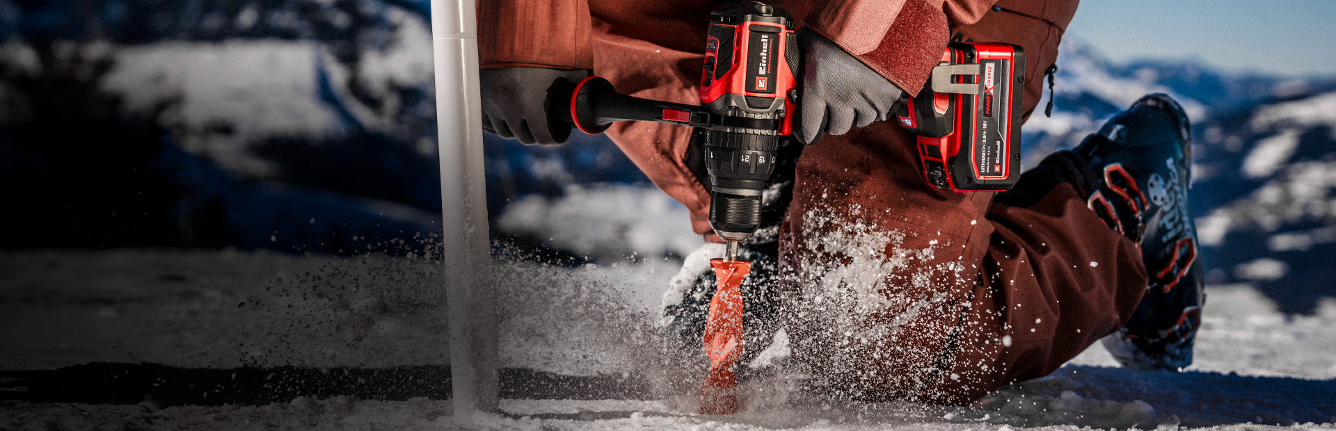 A person wearing winter clothing drills a hole into ice or compacted snow using a cordless power drill, with snow spraying around the drill bit in an outdoor, snowy environment.