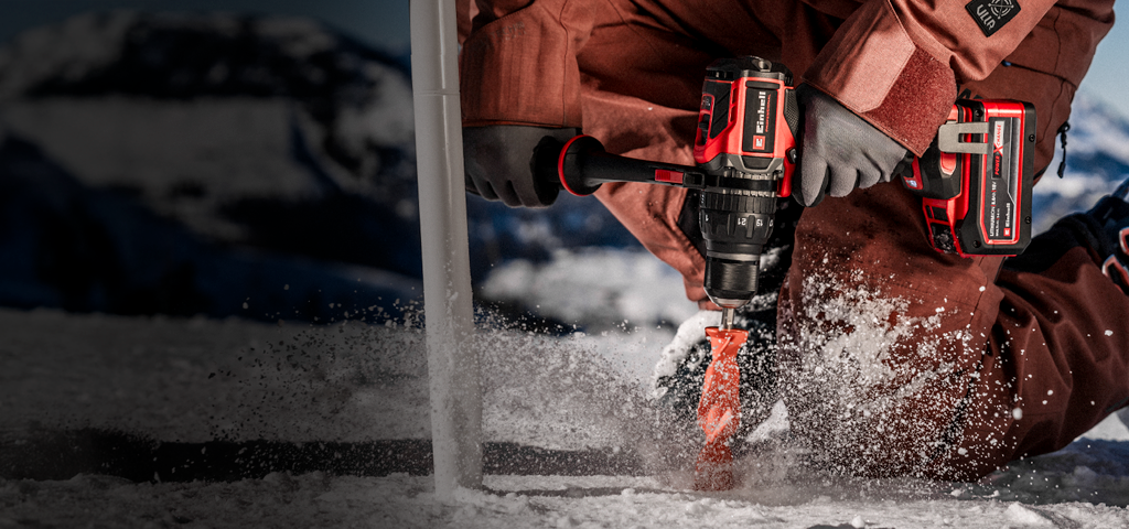 A person wearing winter clothing drills a hole into ice or compacted snow using a cordless power drill, with snow spraying around the drill bit in an outdoor, snowy environment.