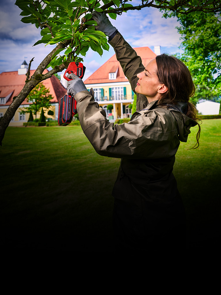 Der Banner zeigt eine Person, die mit einer Akku-Astschere von Einhell einen Baumast schneidet. Die Szene spielt in einem grünen Garten mit einem großen Haus im Hintergrund und hellem Tageslicht. Der Fokus liegt auf der Gartenarbeit und dem verwendeten Gerät.