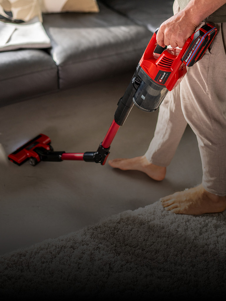 Eine Person saugt den Boden mit einem roten kabellosen Stielstaubsauger von Einhell. Zu sehen ist nur der untere Teil ihres Körpers, und sie steht barfuß in einem modernen Wohnzimmer. Im Hintergrund befindet sich ein graues Sofa mit einer locker darübergelegten Decke.